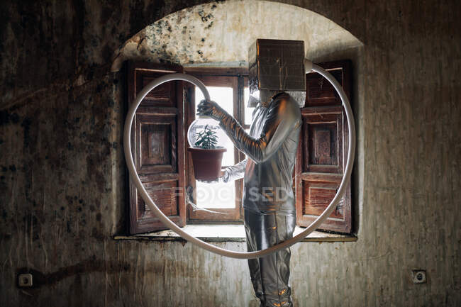 Side view unrecognizable person wearing silver costume with breathing apparatus and hose attached to potted plant standing in shabby abandoned room — Stock Photo