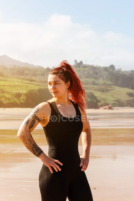Atleta feminina auto-assegurada com as mãos nos quadris olhando para longe na praia do oceano arenoso à luz do sol — Fotografia de Stock