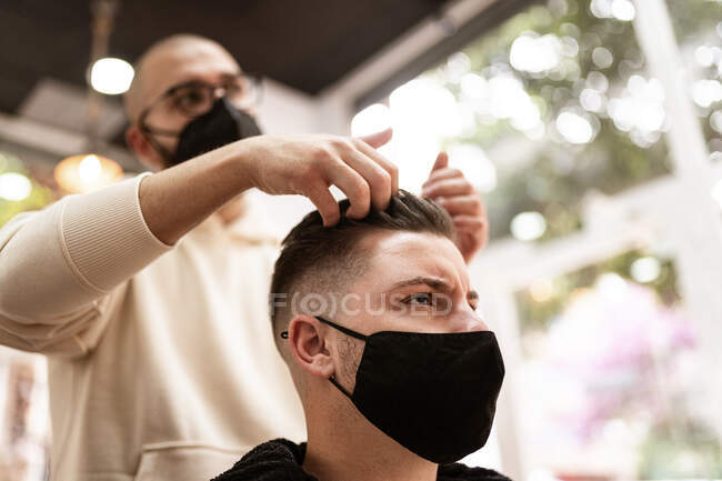 Male hairstylist in eyewear touching hair of client in fabric mask while looking forward in barbershop — Fotografia de Stock