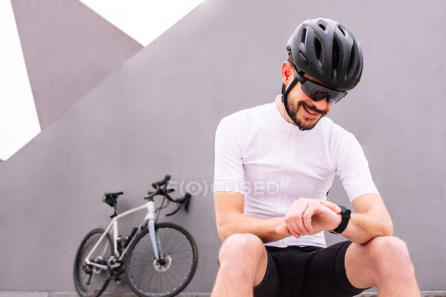 Fröhliche männliche Biker mit Schutzhelm und Sonnenbrille beobachten Herzfrequenz auf tragbarem Armband, während sie gegen Fahrrad auf dreifarbigem Hintergrund sitzen — Stockfoto