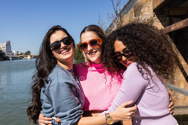 Cheerful multiethnic female friends in casual outfits embracing each other on city waterfront and looking at camera with smiles on sunny summer day — Stock Photo