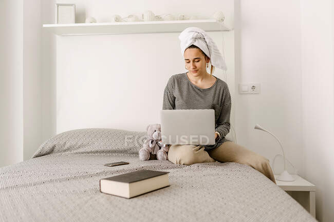 Confident young ethnic female freelancer in casual clothes and towel on head working remotely on netbook while sitting on comfortable bed at home — Fotografia de Stock