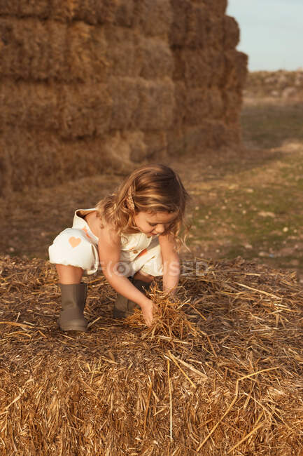 Adorable child in overalls playing with hay near straw bales in countryside — Stock Photo