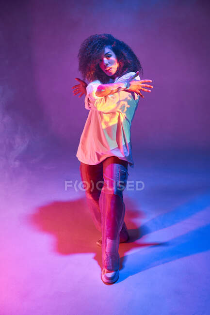 Pleine longueur ajustement jeune danseuse afro-américaine en tenue informelle lâche regardant la caméra tout en dansant en studio sombre dans les néons — Photo de stock