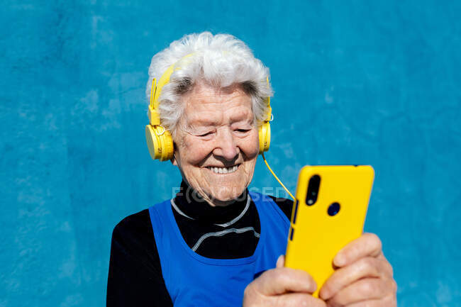 Радостная пожилая женщина в желтых наушниках, слушающая музыку во время просмотра мобильного телефона на ярком синем фоне — стоковое фото
