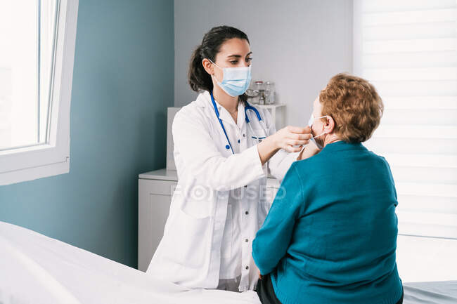 Doctora joven en uniforme que se pone mascarilla desechable en anciana anónima en examen en el hospital durante el brote covid 19 - foto de stock