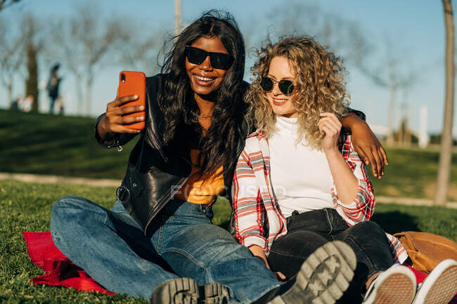 Fröhliche multirassische beste Freundinnen, die sich im Frühlingsgarten umarmen und an sonnigen Tagen Selbstaufnahmen mit dem Smartphone machen — Stockfoto