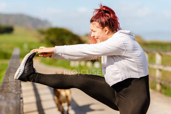 Side view of smiling female athlete in activewear exercising on fenced footpath while looking away on sunny day — Stock Photo