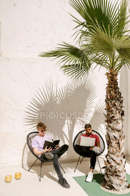Full body of stylish young multiracial male friends using laptop and reading book while relaxing on chairs on terrace on sunny day — Stock Photo