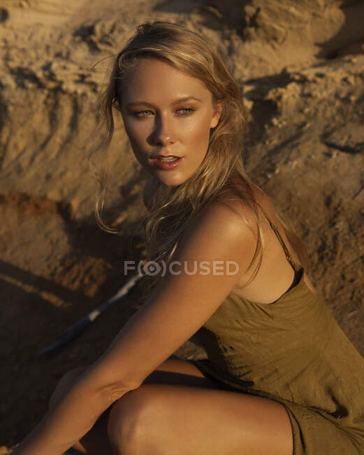 Crop of barefoot sensual blond female sitting on haunches near sandy slope and looking away — Stock Photo