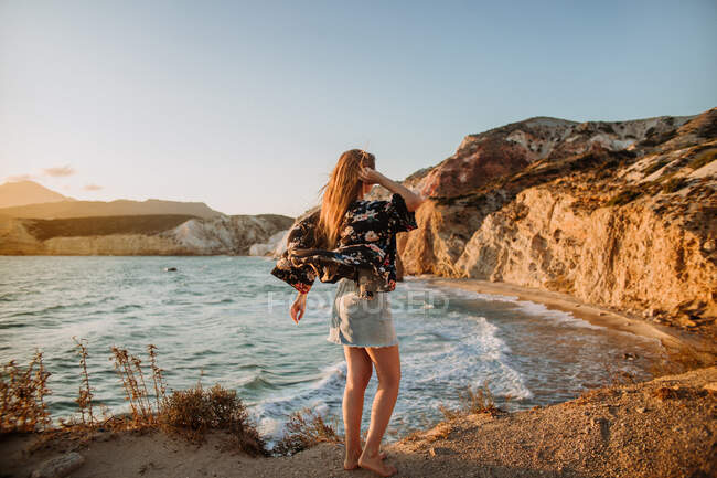 Вид сзади Анонимная женщина в мини-юбке стоит на грубом скалистом берегу моря и трогает длинные волосы под ясным голубым небом в Fyriplaka Милош — стоковое фото