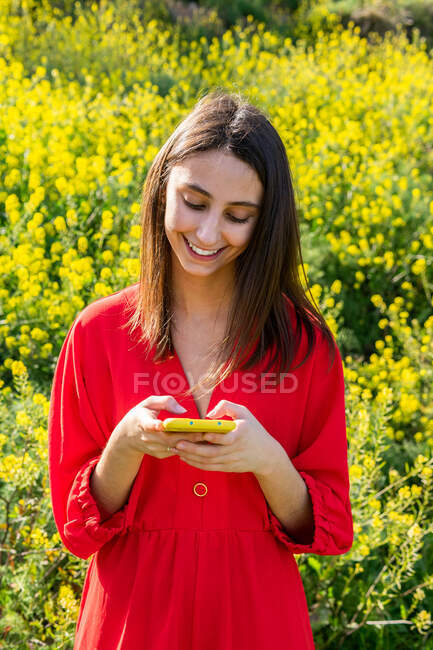 Young content female in red apparel text messaging on cellphone against blossoming plants in sunlight — Stock Photo