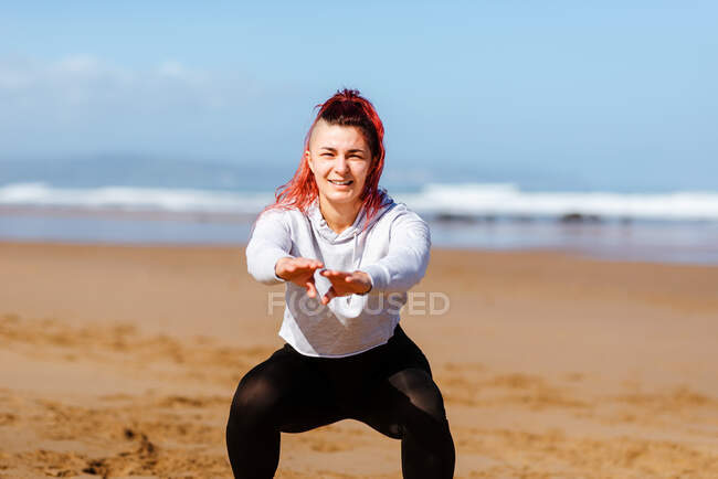 Alegre atleta femenina haciendo ejercicio con los brazos extendidos en la costa del océano arenoso mientras mira la cámara - foto de stock
