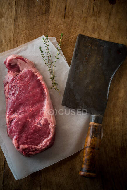 Вид сверху на непрожаренный кусок мяса с черным перцем на бумагу для выпечки и топорик на деревянном фоне — стоковое фото