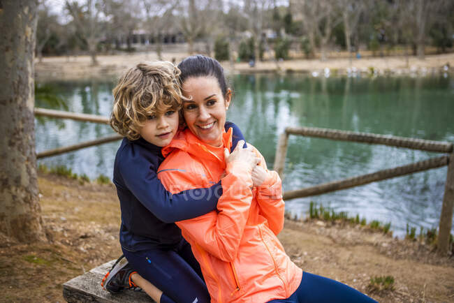 Charming boy embracing smiling mother while sitting and looking away against water in daylight — Stock Photo