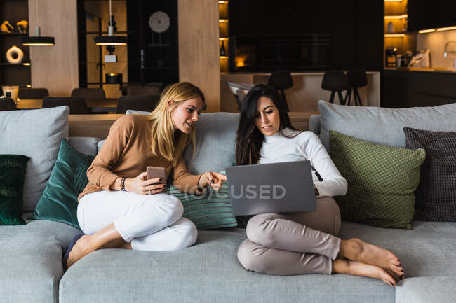 Paar fröhlicher homosexueller Frauen, die auf der Couch chillen und Videos auf dem Smartphone ansehen und am Laptop arbeiten, während sie am Wochenende zusammen unterhalten — Stockfoto