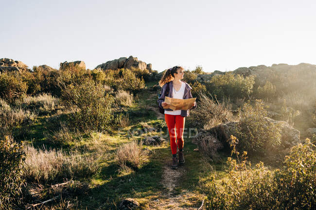 Повне тіло зосередженої молодої жінки-дослідника в повсякденному одязі, що читає карту під час прогулянки на схилі гори в сонячний день — стокове фото
