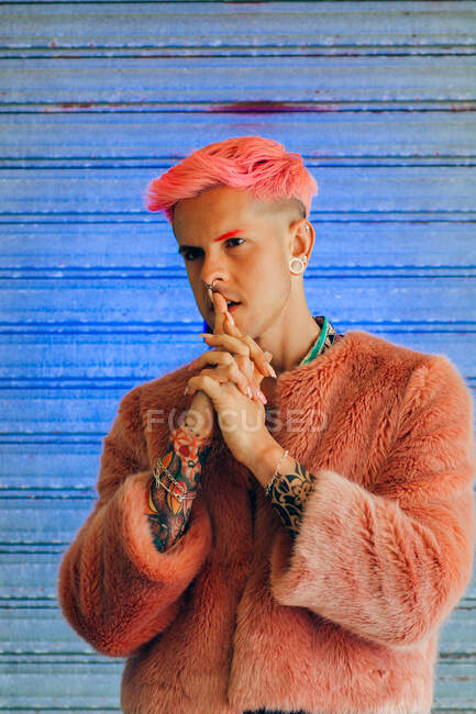 Hombre homosexual joven pensativo en ropa de moda con manicura y corte de pelo moderno mirando hacia otro lado sobre fondo azul - foto de stock