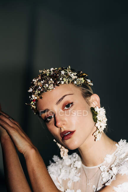 Charmante zarte junge Braut in weißem Spitzenkleid und luxuriösem Blumenkranz und Ohrringen blickt vor schwarzem Hintergrund in die Kamera — Stockfoto