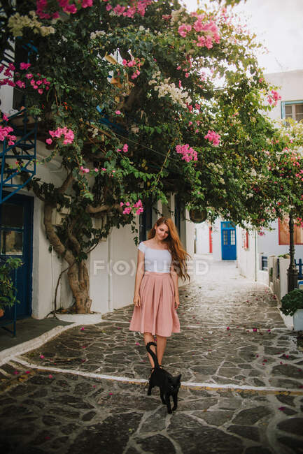 Femme pleine longueur en robe élégante touchant fleurs roses arbre en fleurs tout en se tenant sur la rue piétonne en pierre près de chat noir dans un petit village authentique en Grèce — Photo de stock