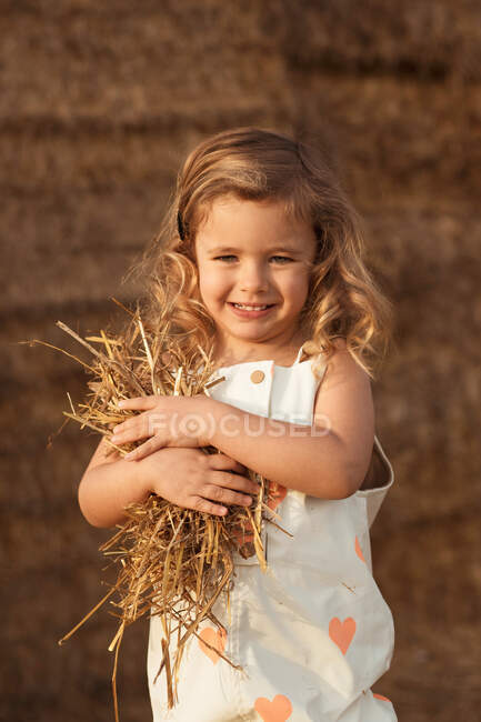 Cheerful adorable child in overalls playing with hay near straw bales in countryside — Stock Photo