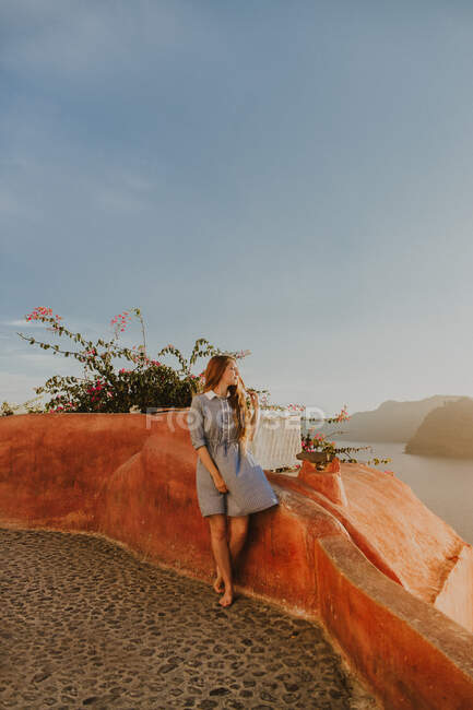 Full length young female in sundress sitting on coastal town embankment railing and admiring picturesque views on sunny day in Santorini — Stock Photo