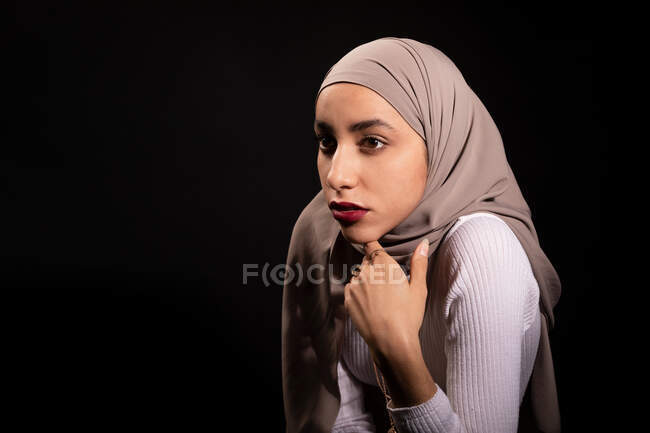 Modish confident Muslim female in hijab leaning on chair and looking away in dark studio — Stock Photo