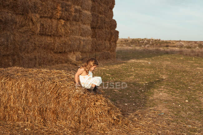 Side view of curious little girl in overalls climbing straw bale while playing in evening in countryside — Stock Photo