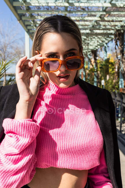 Atractiva joven hembra en jeans suéter rosa y abrigo negro de pie con las manos en los bolsillos cerca de la mesa en el restaurante y mirando a la cámara - foto de stock