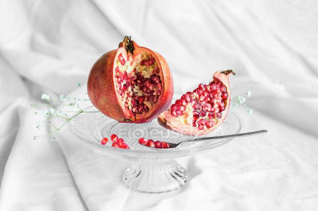 Bright tasty fresh pomegranate with ripe seeds and blooming flower sprig on transparent stand on crumpled fabric — Fotografia de Stock