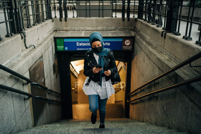 Ethnic female in hijab and protective mask walking upstairs in city street while using smartphone — Fotografia de Stock