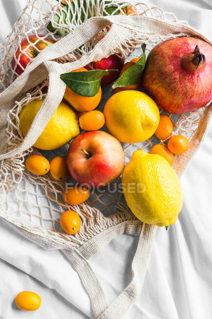From above of different whole ripe apple with lemons near pomegranate and kumquats in eco bag on creased cloth - foto de stock