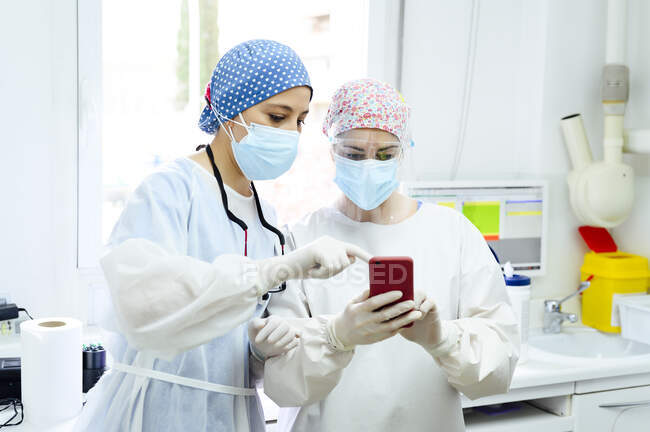 Cirujanos femeninos en gorras de tela médica navegando por Internet en el teléfono celular contra la computadora de escritorio en el hospital ligero - foto de stock