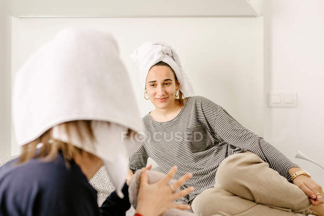 Happy young ethnic female best friends in casual clothes and towels on heads relaxing on comfortable bed after having bath at home — Fotografia de Stock