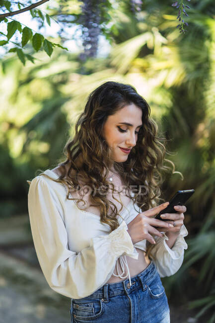 Привабливий контент жінки в повсякденному одязі з голим переглядом живота мобільний телефон у багатому парку на ясну теплу погоду — стокове фото