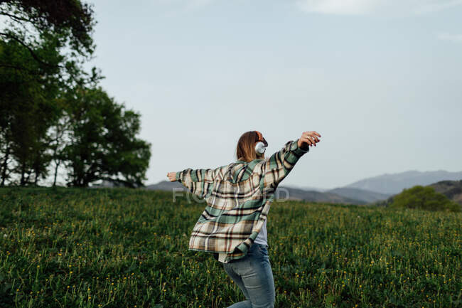 Rückansicht einer anonymen Frau, die über Kopfhörer Musik hört, während sie sich auf der Sommerwiese vor den Bergen vergnügt — Stockfoto