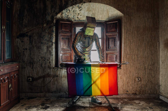 Неузнаваемый человек в серебряном костюме и коробке на голове гладит флаг ЛГБТК, стоя в убогой заброшенной комнате — стоковое фото