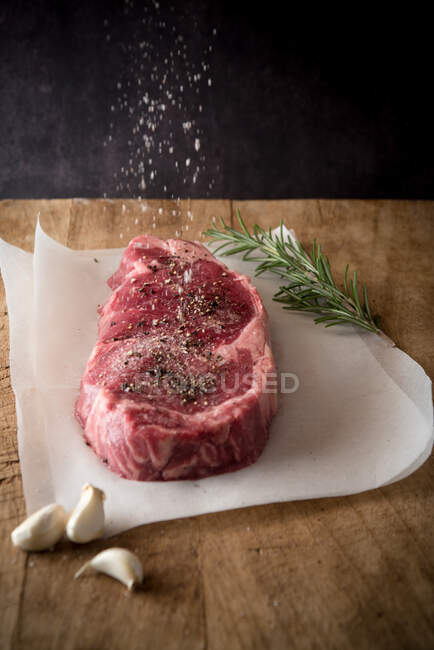 Pedaço de carne crua com pimenta preta moída sob sal no ar entre dentes de alho frescos e raminho de alecrim — Fotografia de Stock