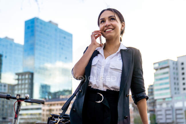 Fröhliche junge ethnische Unternehmerin telefoniert, während sie im Sonnenlicht gegen den Stadtfluss schaut — Stockfoto