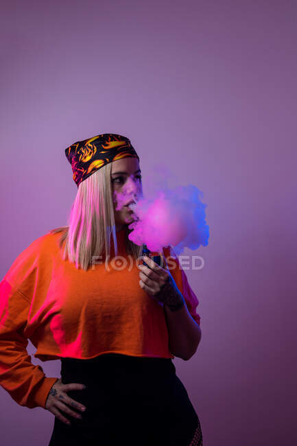 Cool female in street style outfit smoking e cigarette and exhaling smoke through nose and mouth on purple background in studio with pink neon illumination — Stock Photo