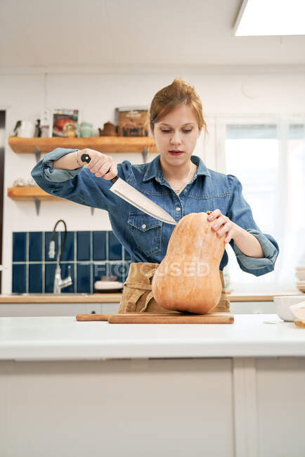 Jeune femme avec couteau tranchant coupant la courge crue sur la planche à découper tout en cuisinant dans la cuisine à la maison — Photo de stock
