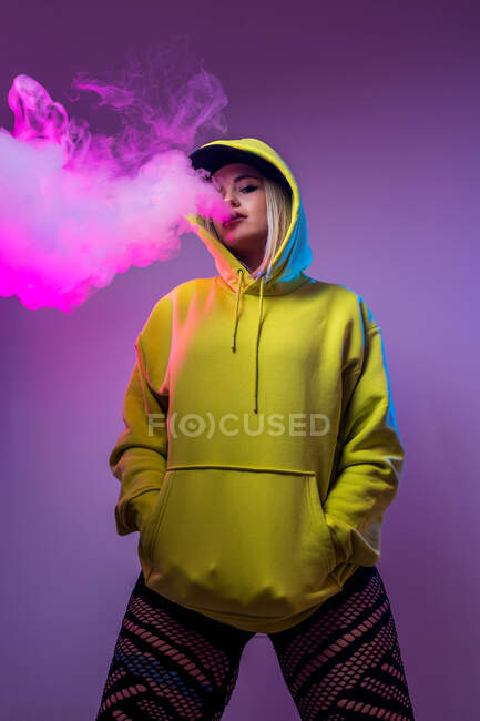 Знизу впевнена жінка-хіпстер у светрі курить електронну сигарету в студії на рожевому фоні і дивиться на камеру — стокове фото