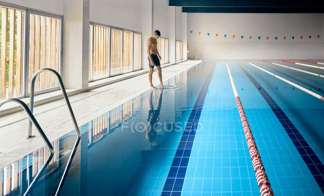 Vista lateral do atleta masculino em calções de banho em pé contra a piscina com pistas e água pura antes do treino — Fotografia de Stock