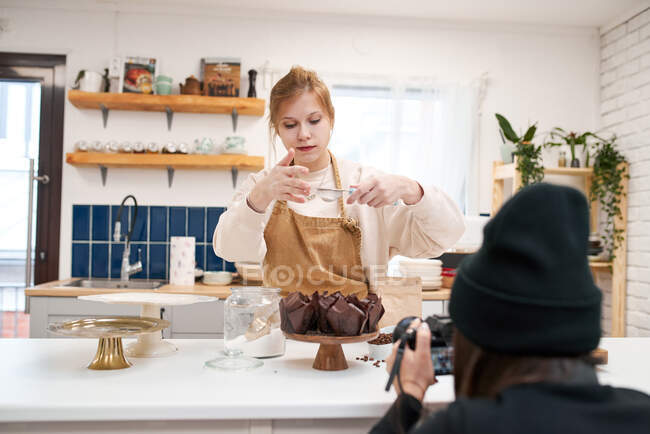 Lächelnde junge Frau mit Sieb bestreut Muffins in Backbecher mit Puderzucker in der Hausküche — Stockfoto