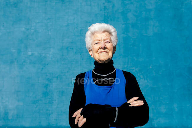 Урожай щасливої зрілої жінки в спортивному одязі, що стоїть на синій стіні у відкритому спортивному центрі і дивиться на камеру з посмішкою і схрещеними руками — стокове фото