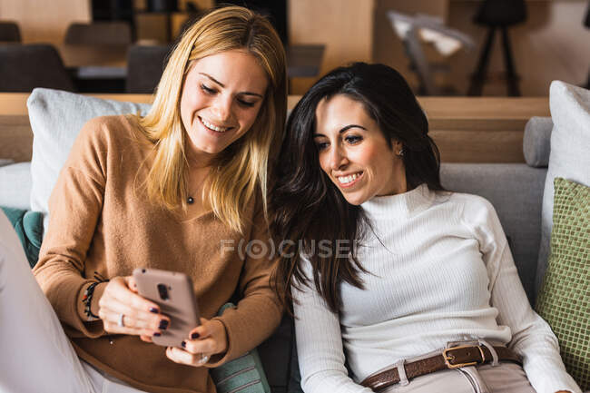 Pareja de mujeres homosexuales sentadas en el sofá y viendo videos divertidos en el teléfono móvil mientras se ríen y se divierten - foto de stock
