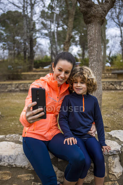 Cheerful mom in sports clothes embracing charming boy while taking self portrait on cellphone in park — Stock Photo