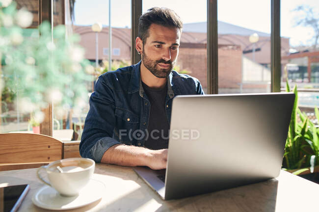 Barba etnica maschio dipendente remoto digitando sul netbook a tavola con una tazza di caffè alla luce del sole — Foto stock
