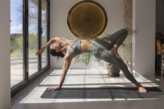 Equilíbrio feminino flexível gracioso em Camatkarasana no tapete enquanto pratica ioga no quarto com luz solar em casa — Fotografia de Stock
