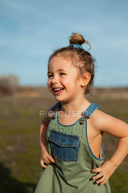 Delighted adorable little girl in overalls standing with hands on waist in meadow and looking away — Stock Photo